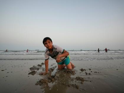 A boy plays in wet sand as others swim and commercial vessels lie anchored in the Strait of Hormuz off Bandar Abbas, Iran.
