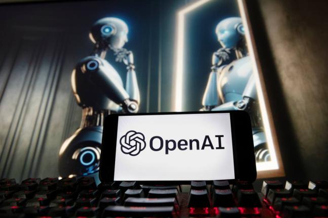 The OpenAI logo is displayed on a cellphone with an image on a computer monitor generated by ChatGPT’s Dall-E text-to-image model.
