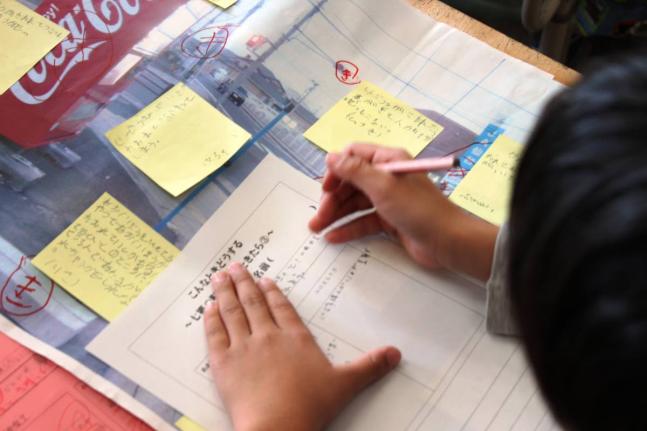 A Grade 2 student of Shichigo Elementary School, Sendai, completes a form on disaster preparedness. On the desk is a poster of the route they take to get home. On the Stick-it notes are the suggestions given by students.