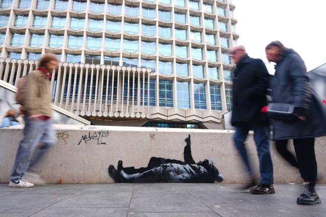 Pedestrians walk past a graffiti artwork, depicting two people, outside Tottenham Court Road Station in London, Monday, December 22, 2025 as elusive street artist Banksy appeared to confirm Monday that an identical image appeared on a wall on the side of a